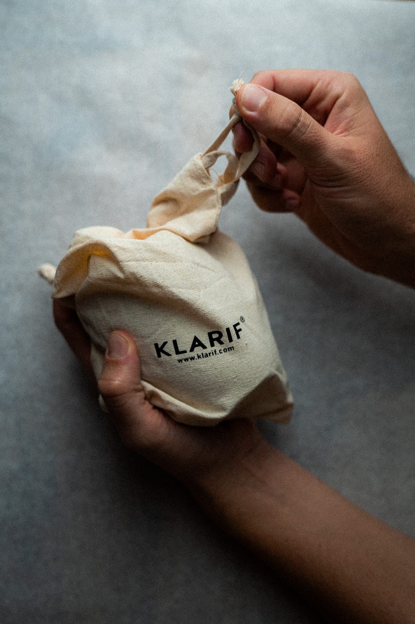 Hands untying a cream cotton drawstring pouch stamped with the Klarif logo in soft, moody studio light