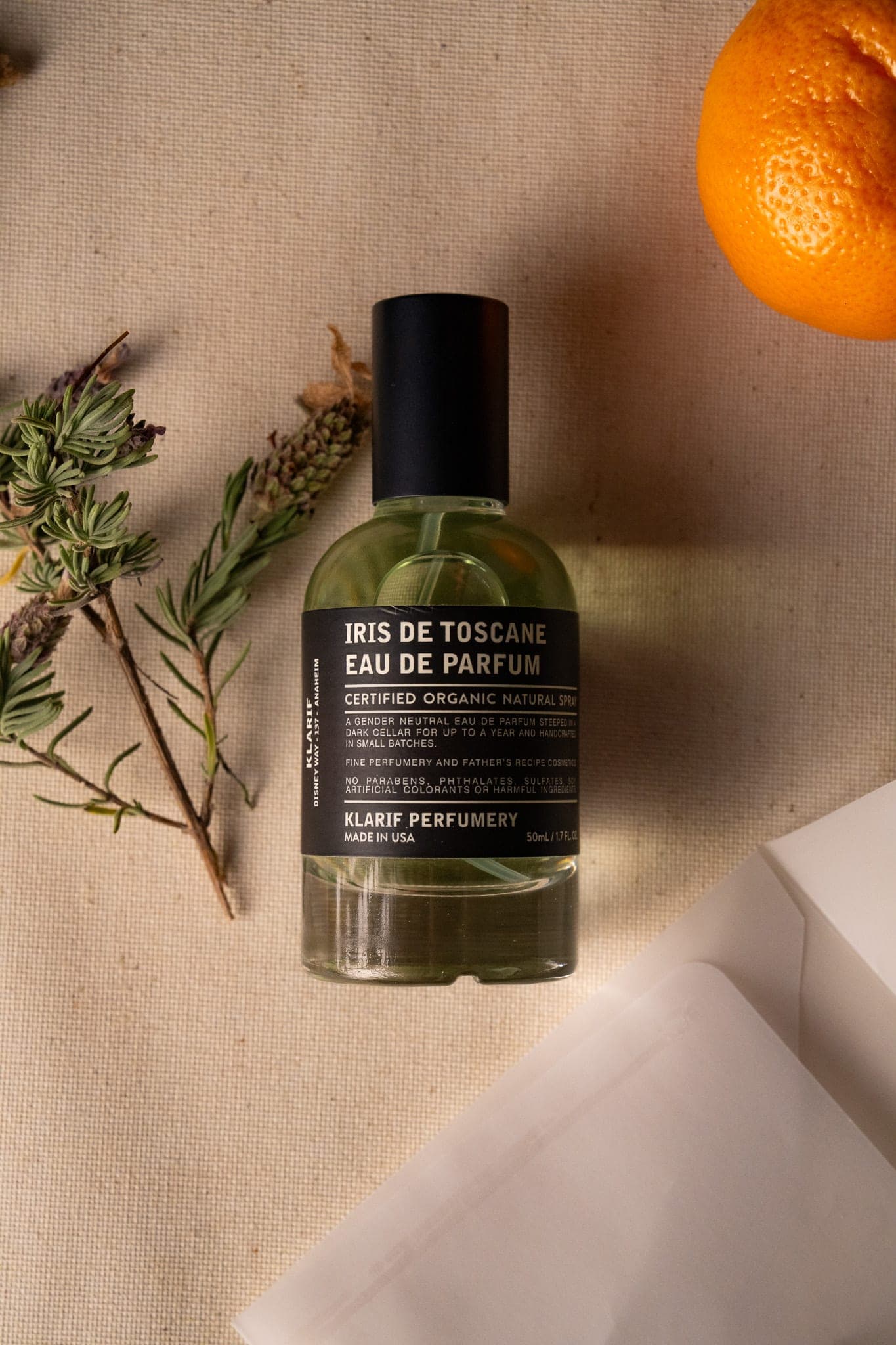 Flat lay of a Klarif eau de parfum bottle beside dried lavender and an orange on warm cream canvas