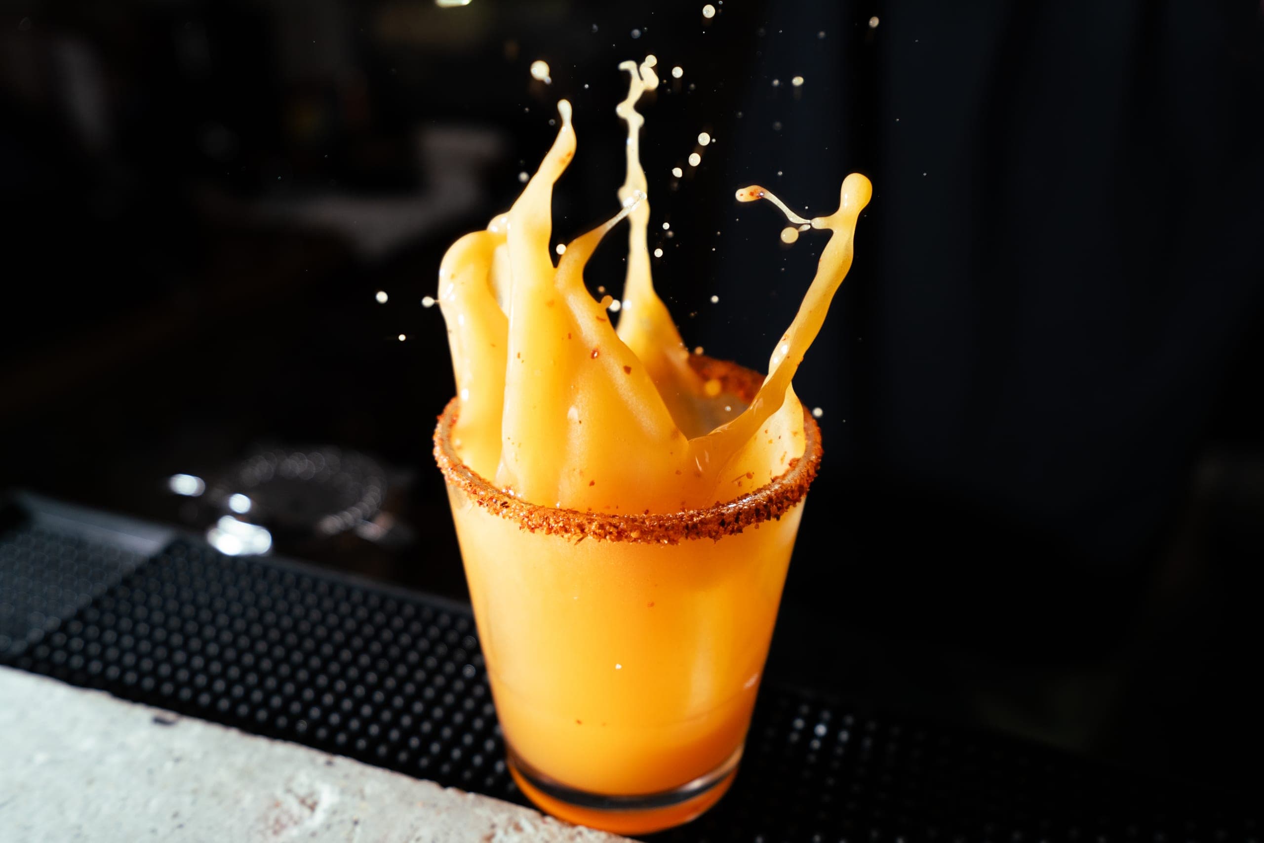 Orange cocktail splashing dramatically out of a chili-salt rimmed glass on a dark bar top