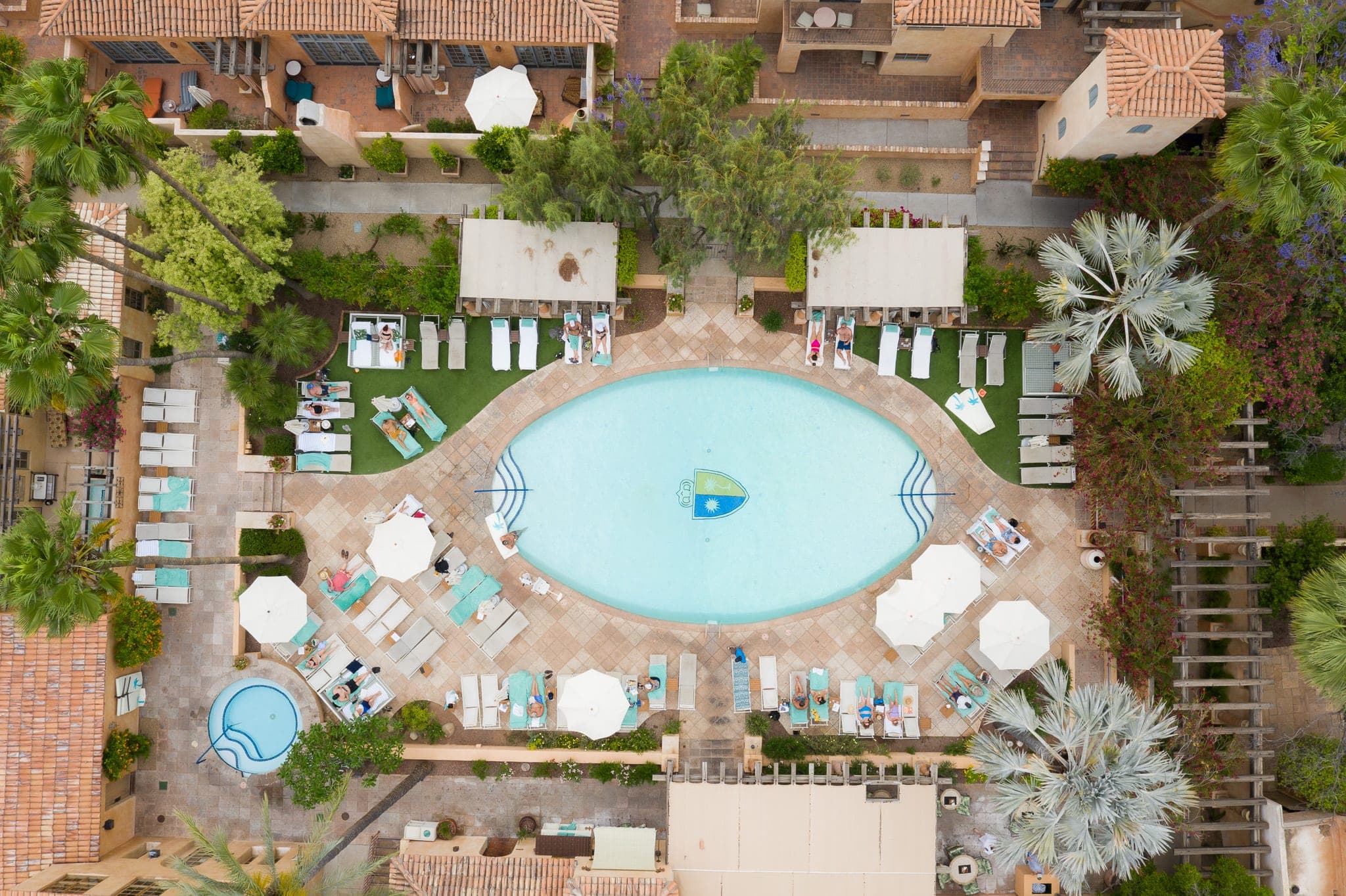 Aerial view straight down over a resort pool ringed by sunbathers, umbrellas, and palms