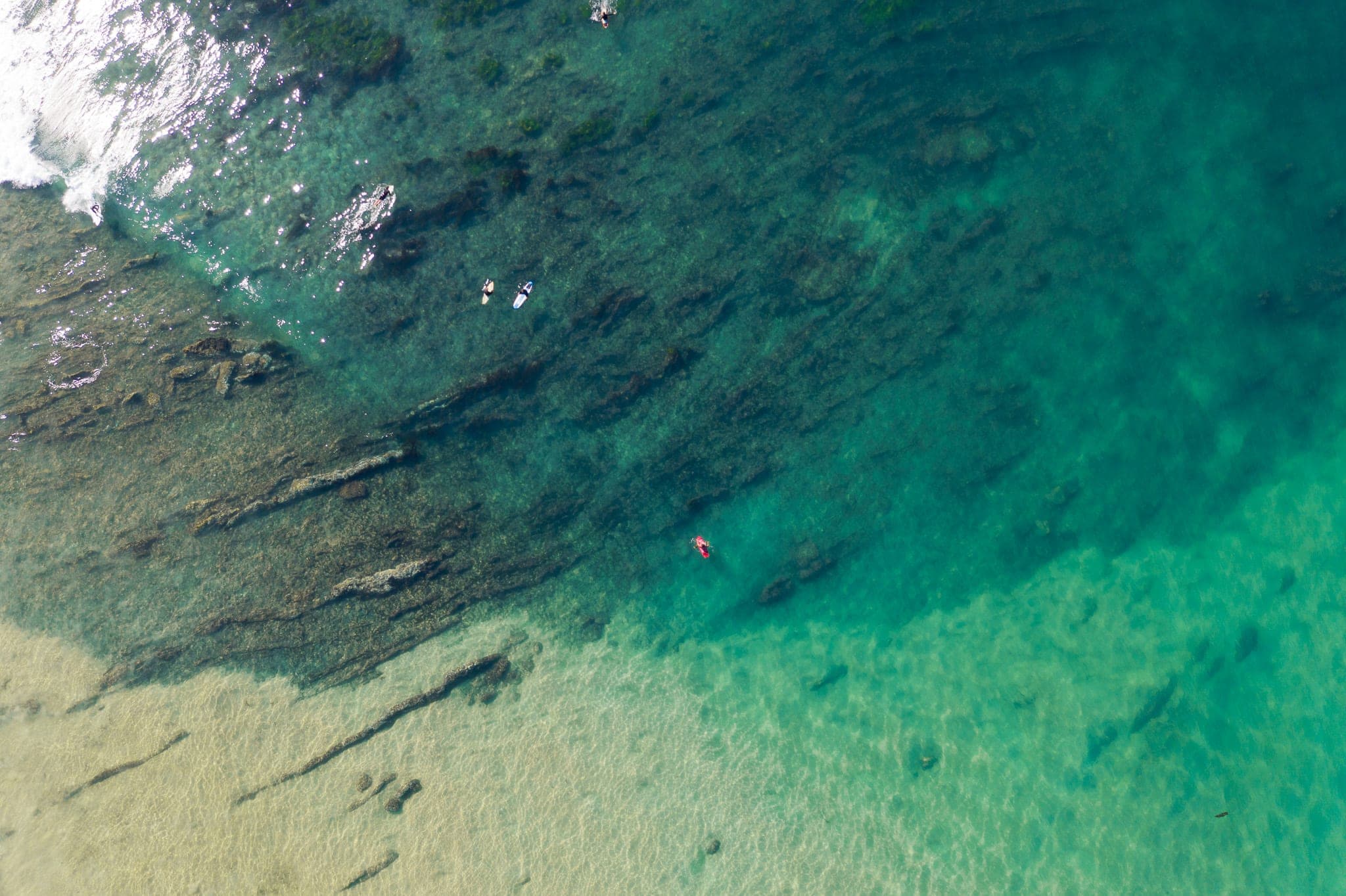 Aerial view of surfers paddling over a rocky reef in gradient turquoise water