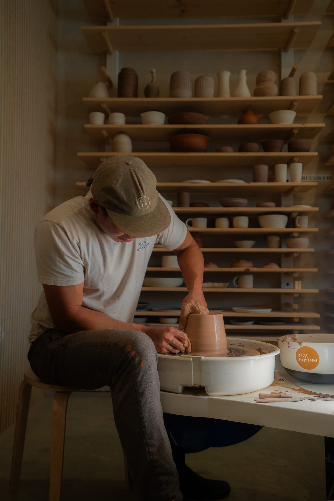 Potter in a ball cap shaping a vessel at the wheel with shelves of unfired pottery behind him