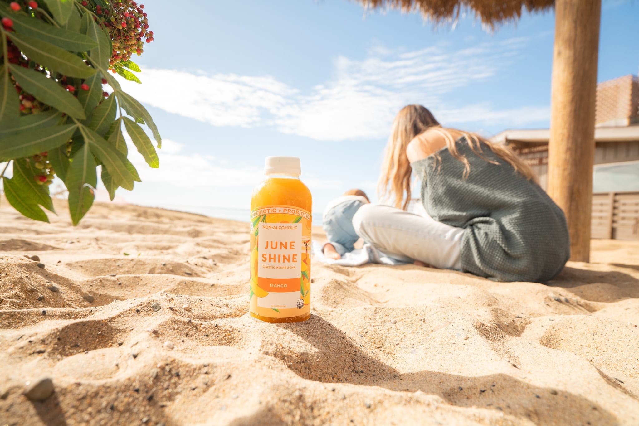 JuneShine mango kombucha bottle standing in beach sand while a woman and toddler relax under a palapa