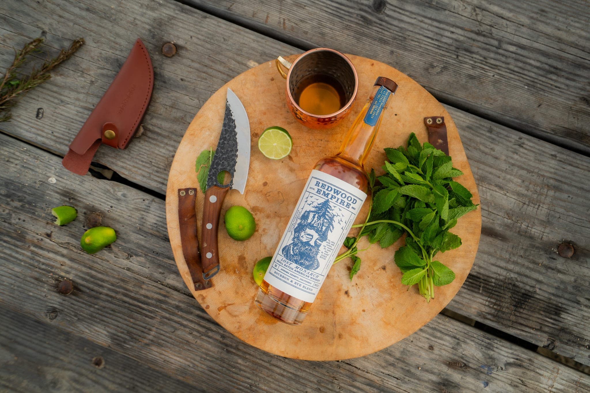 Whiskey bottle styled on a round cutting board with mint, limes, a knife and a copper mug on weathered planks