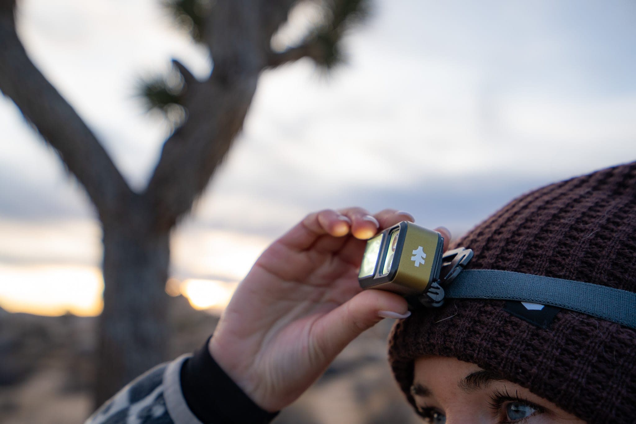 Close-up of a woman adjusting a glowing headlamp on her beanie at dusk in the desert
