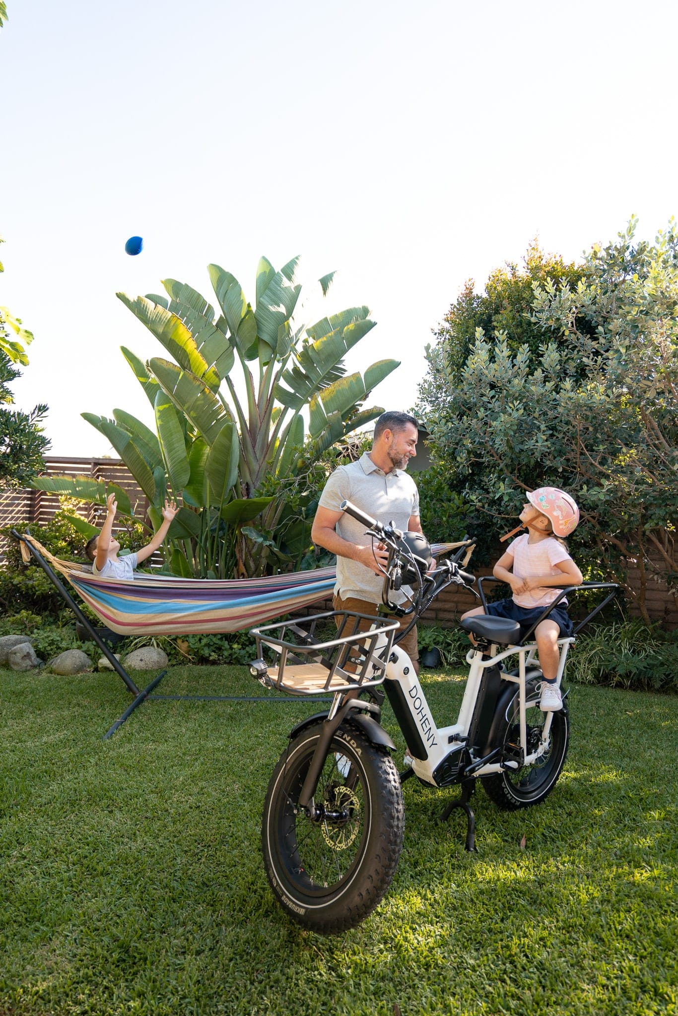 Father and daughter with a cargo e-bike in a lush backyard while a boy plays in a hammock