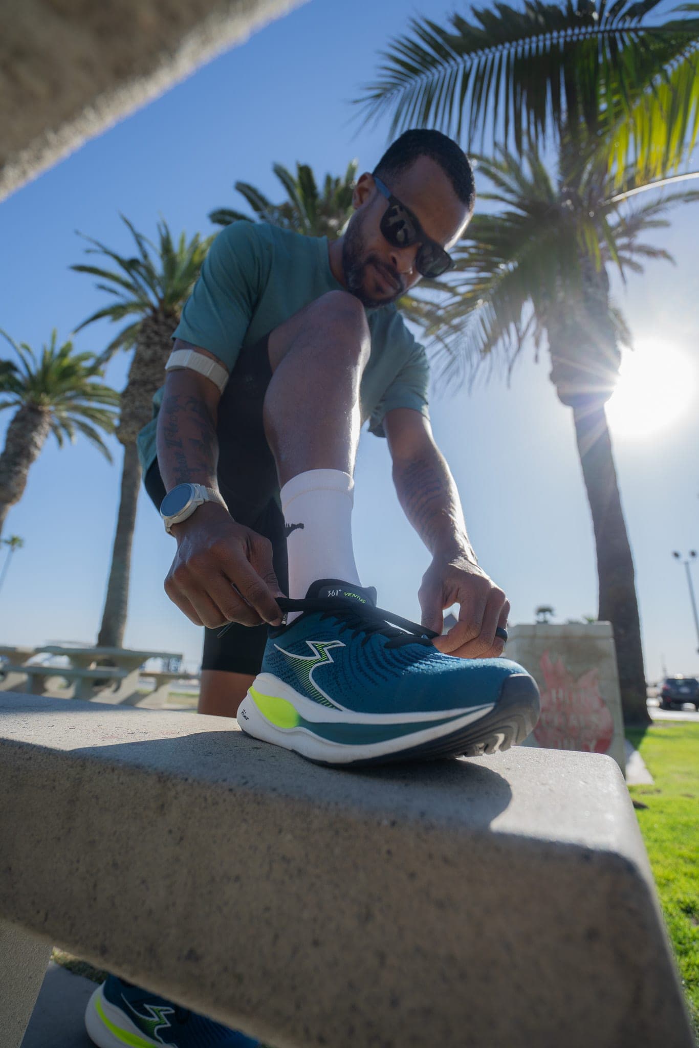Runner lacing up a blue running shoe on a concrete bench beneath backlit palm trees