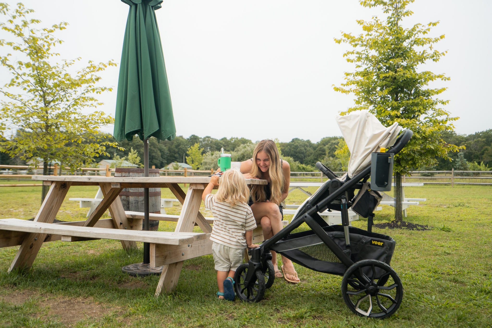 Mother leaning across a picnic table to her toddler beside a parked stroller on a grassy patio