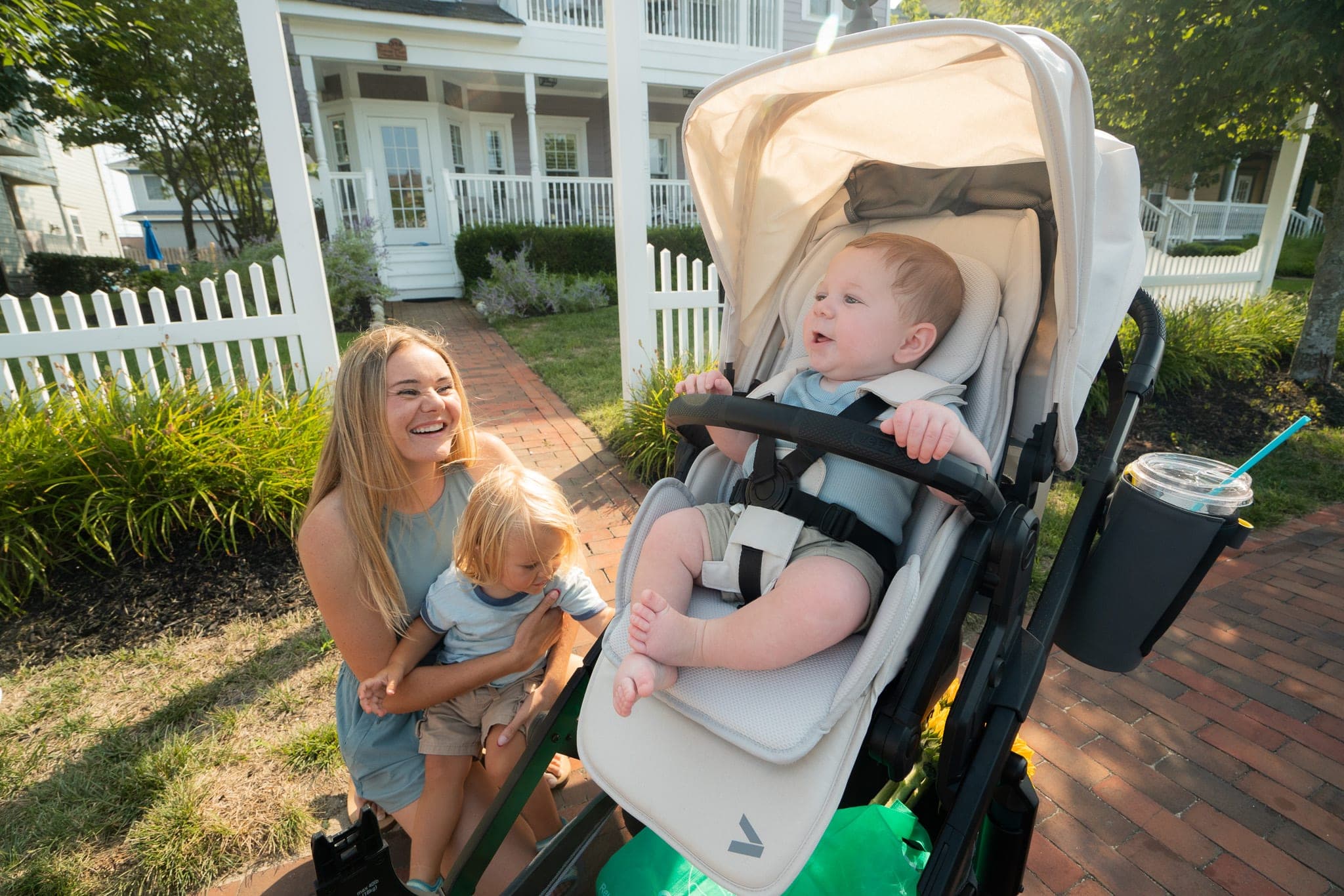 Laughing mother kneeling with her toddler next to a baby strapped into a stroller in front of a picket-fenced house