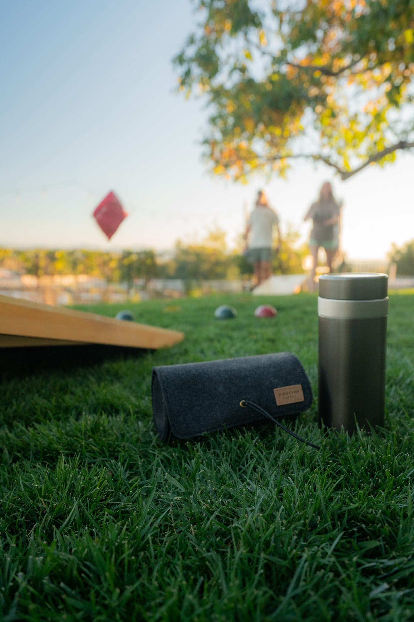 Insulated flask and felt pouch on a lawn while friends toss cornhole bags in the sunset background