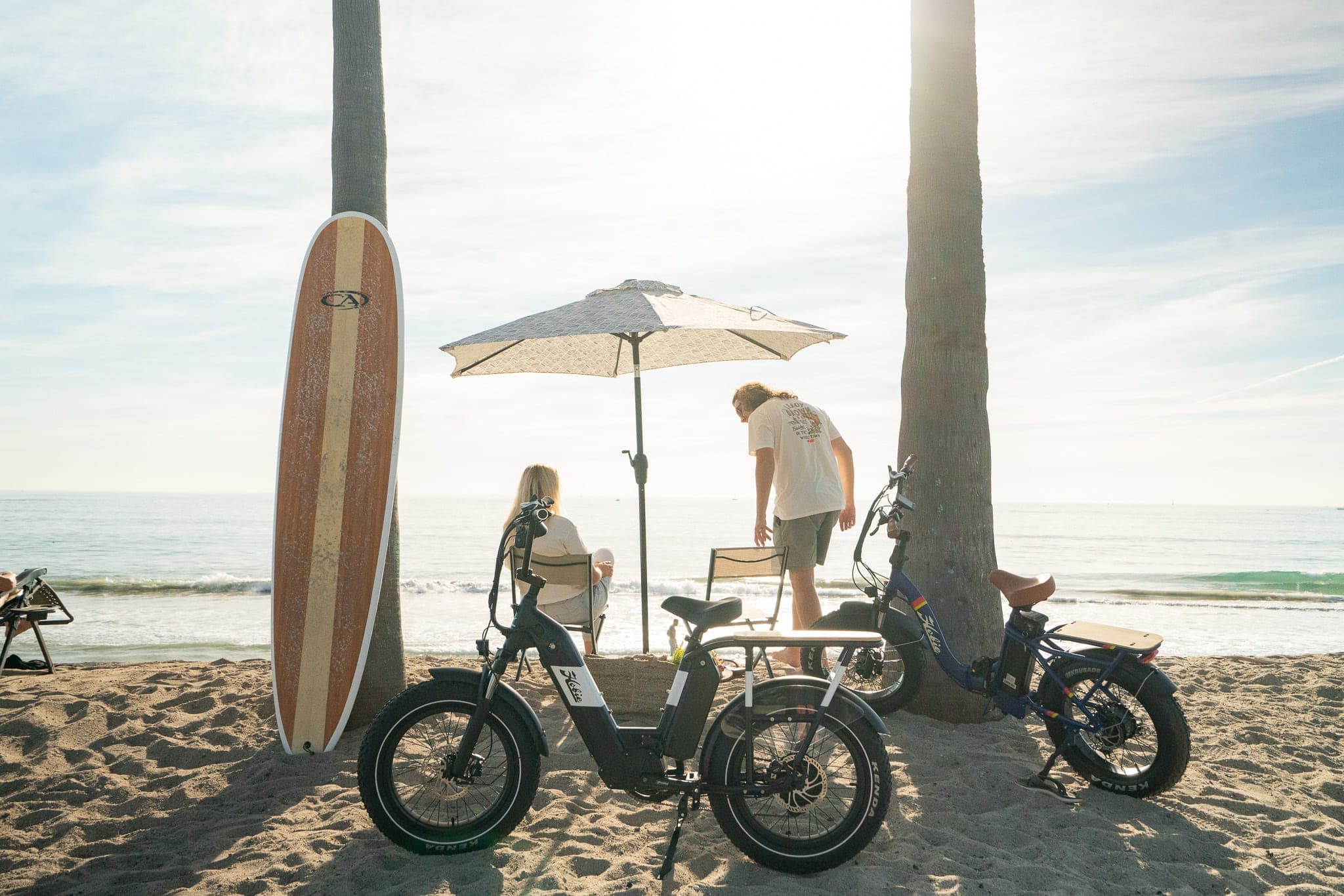 Couple relaxing under a beach umbrella between palm trees with electric bikes and a longboard in golden light