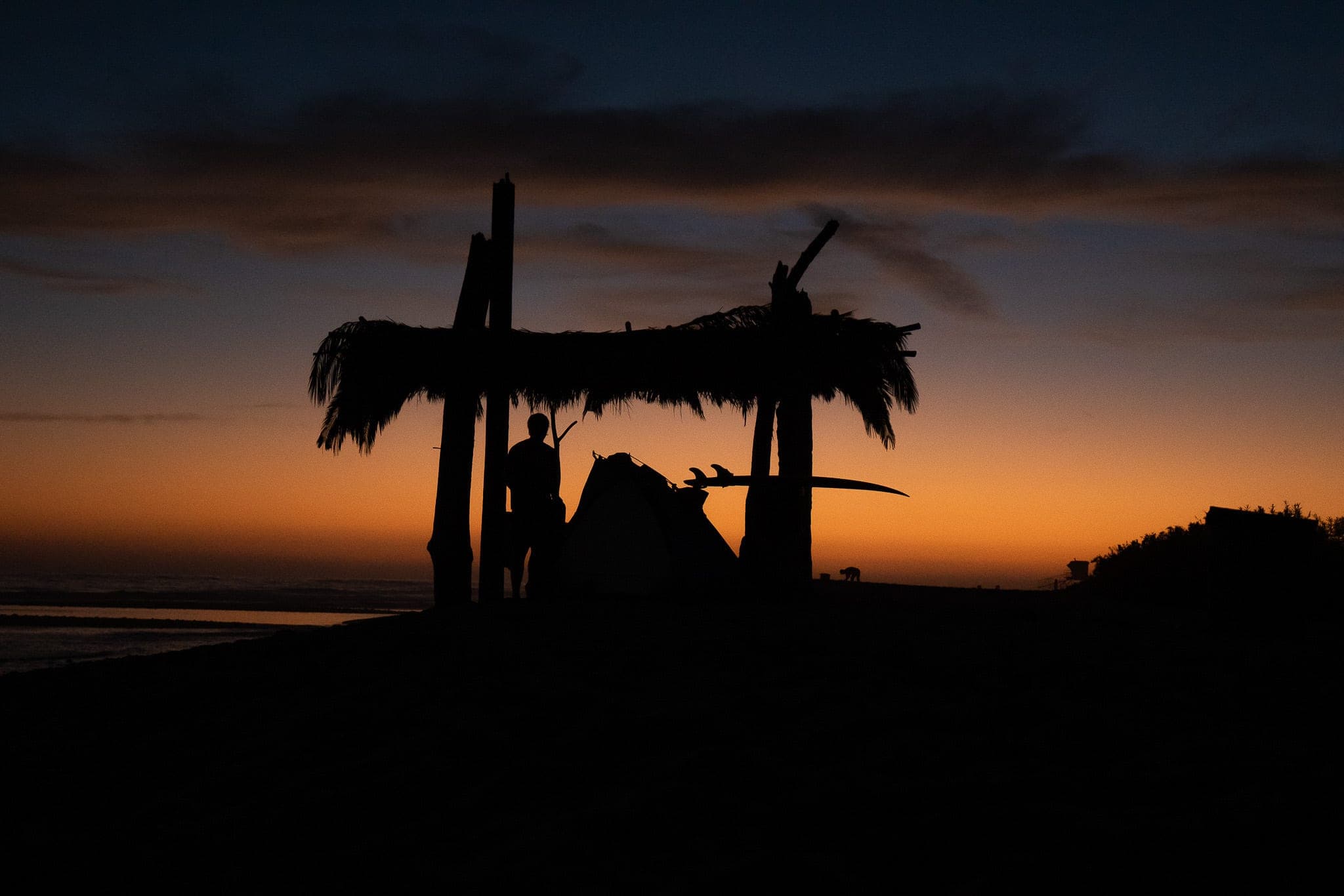 Silhouette of a surfer and palm-thatched palapa against a deep orange sunset sky on the beach