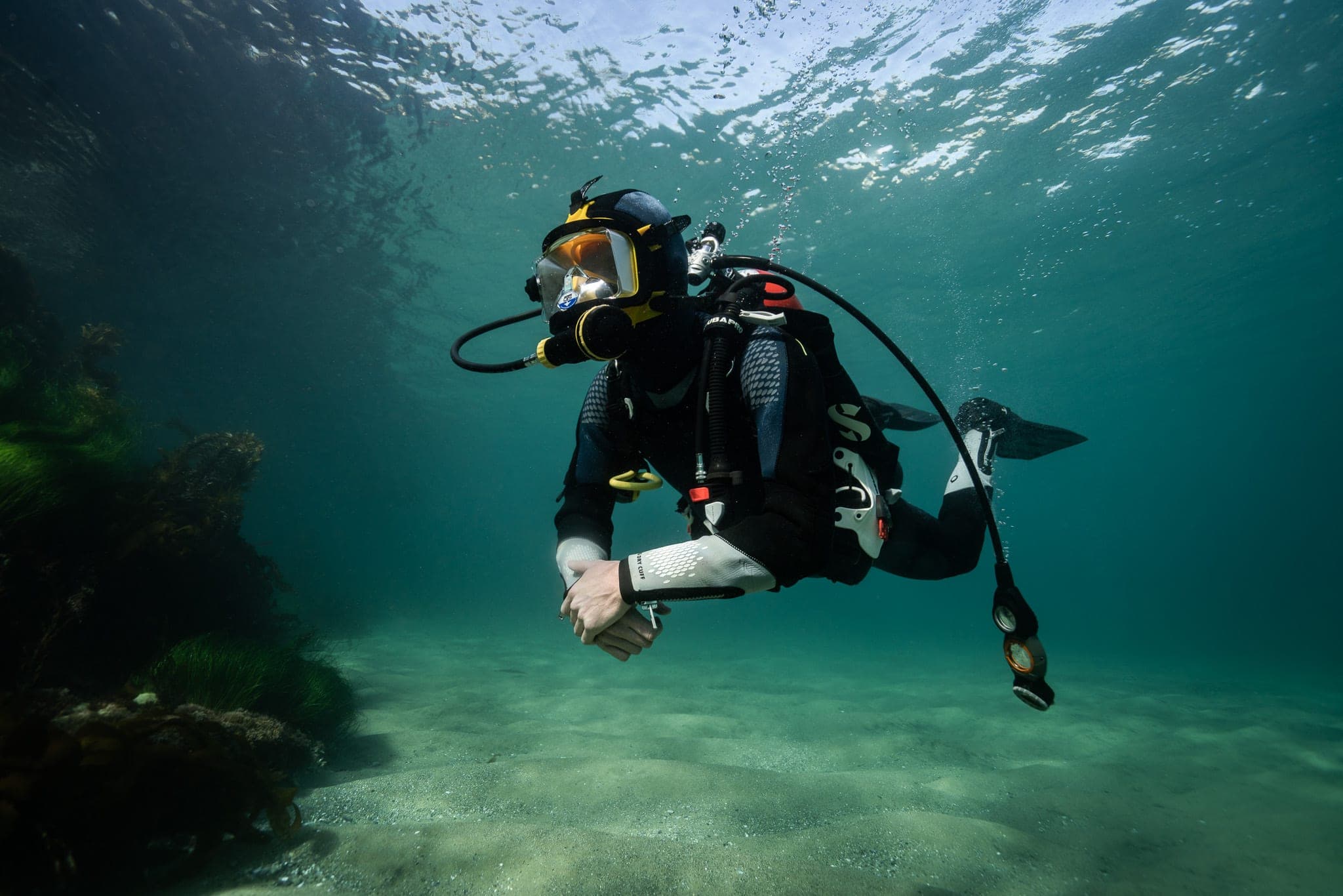 Scuba diver in a full-face mask gliding over a sandy seafloor beside kelp in teal water