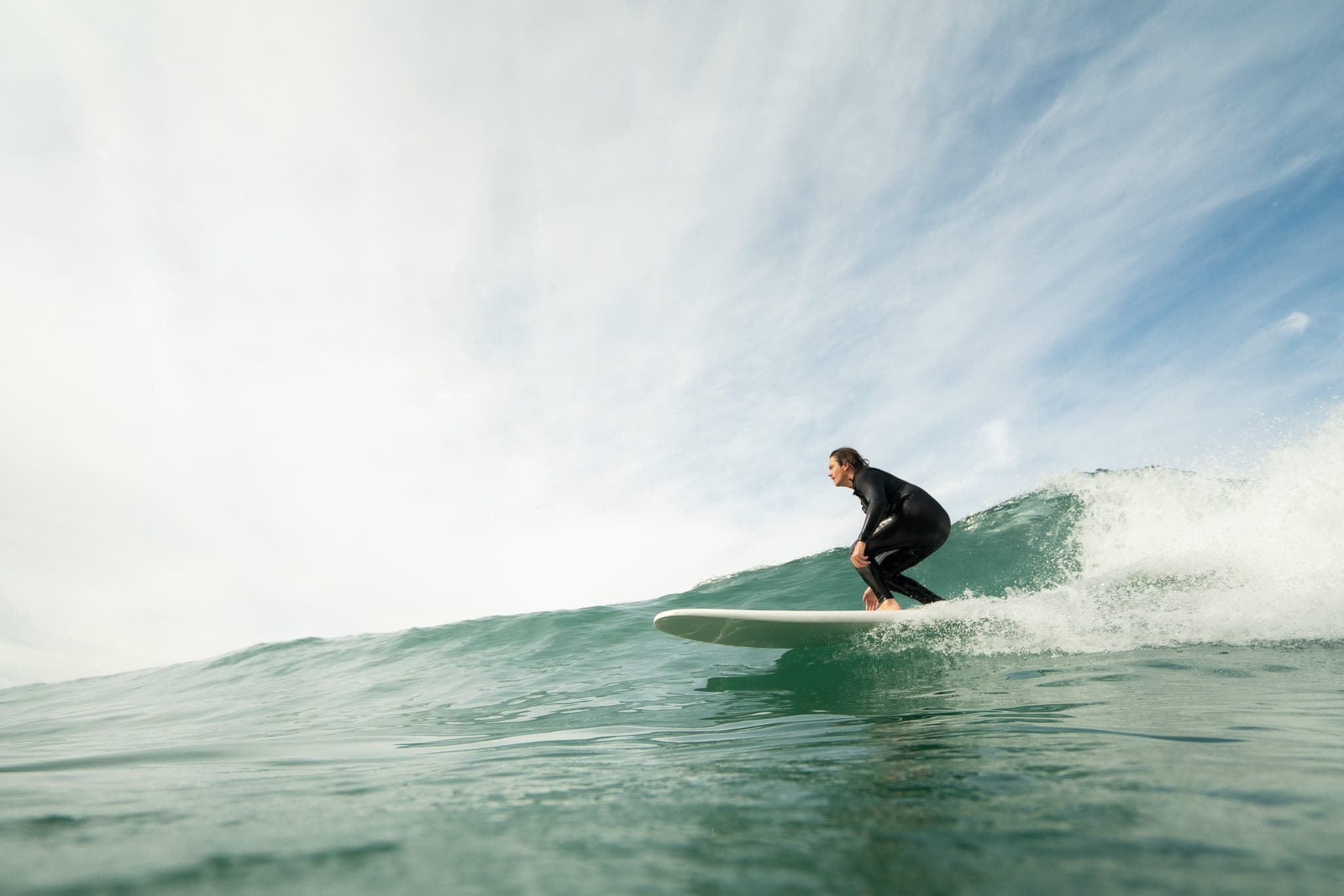 Surfer in a black wetsuit crouching on a longboard as a small green wave peels behind him
