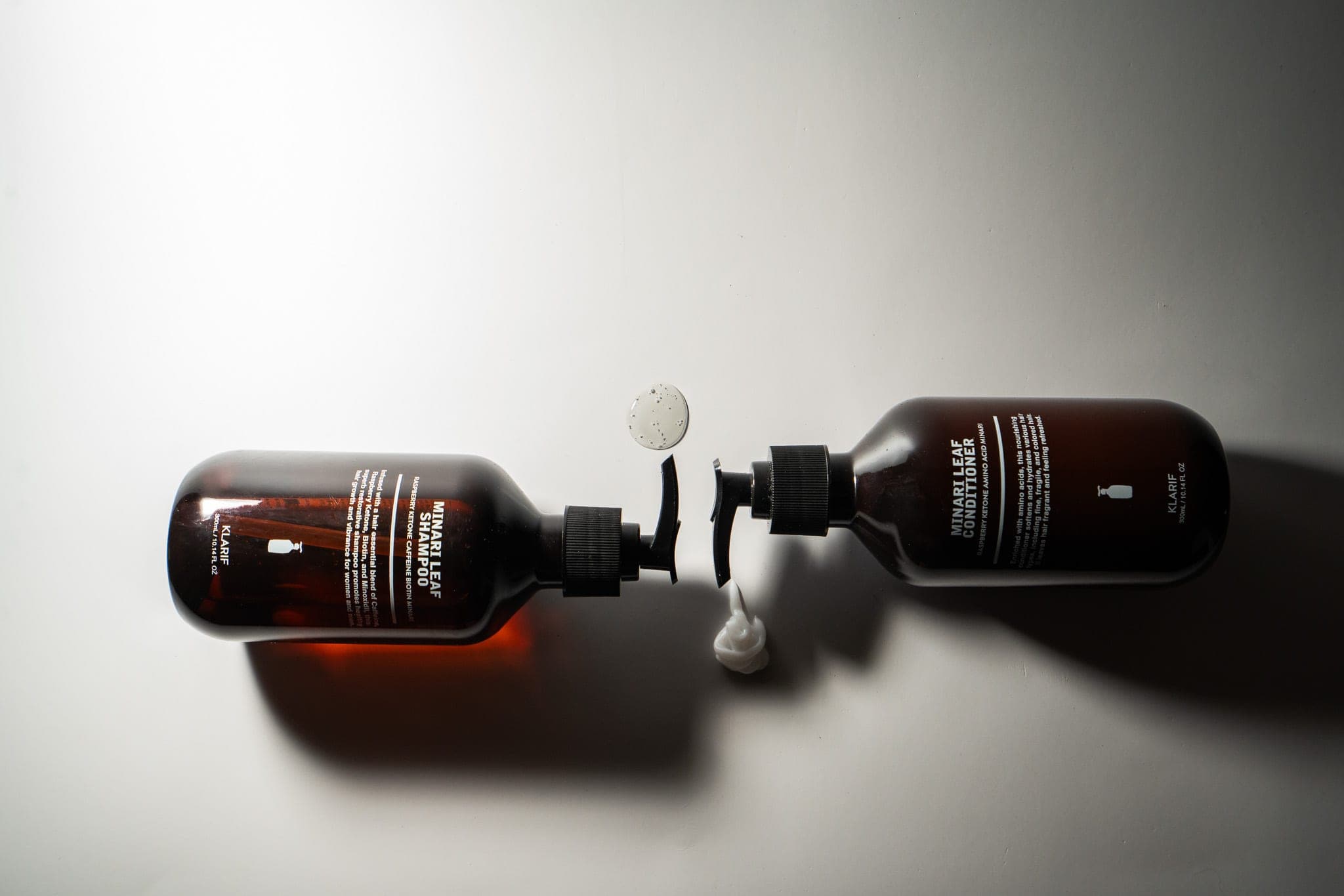 Amber shampoo and conditioner bottles lying head-to-head on a grey backdrop with product swatches between them