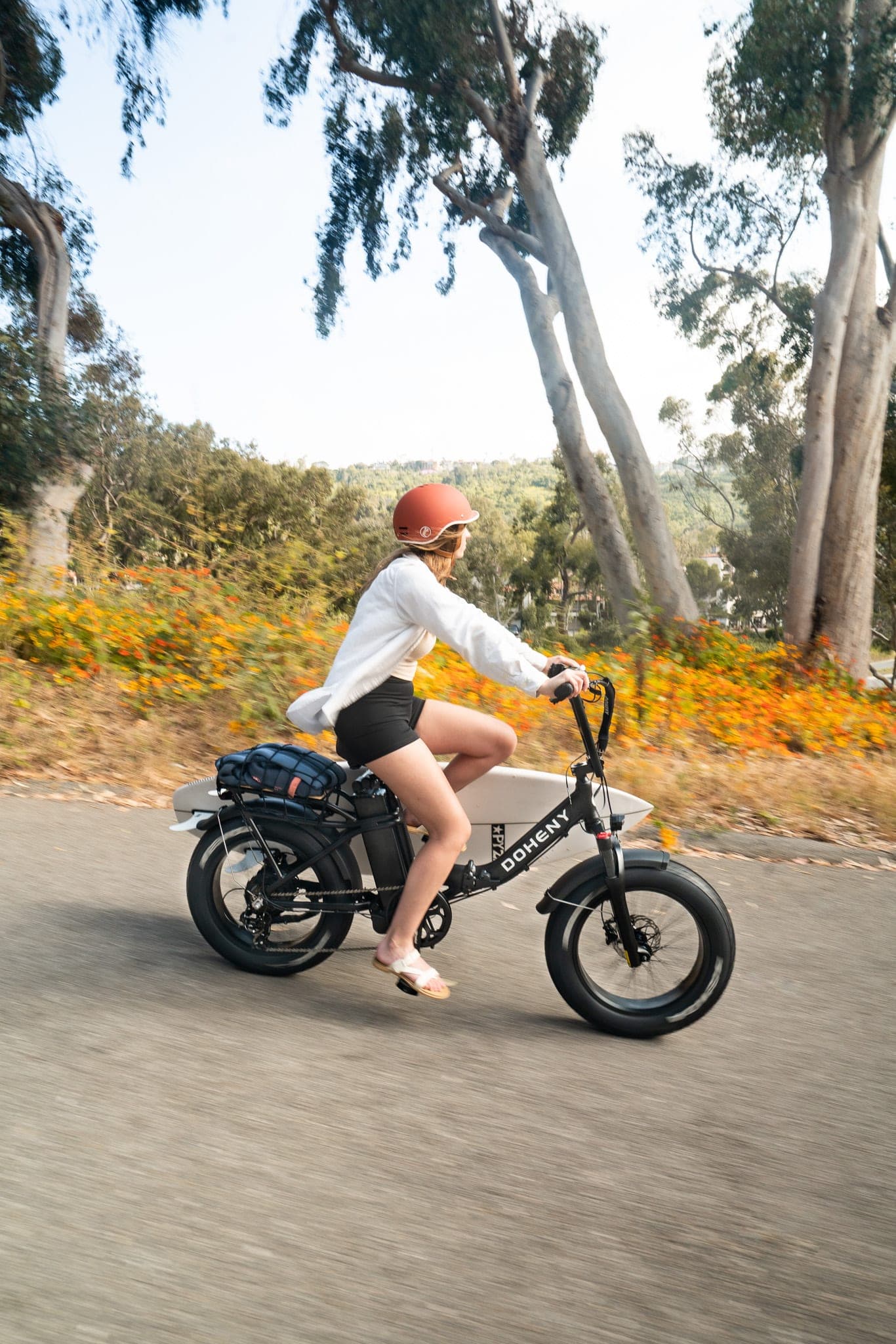 Woman in a red helmet riding an electric bike with a surfboard rack past eucalyptus trees and orange wildflowers