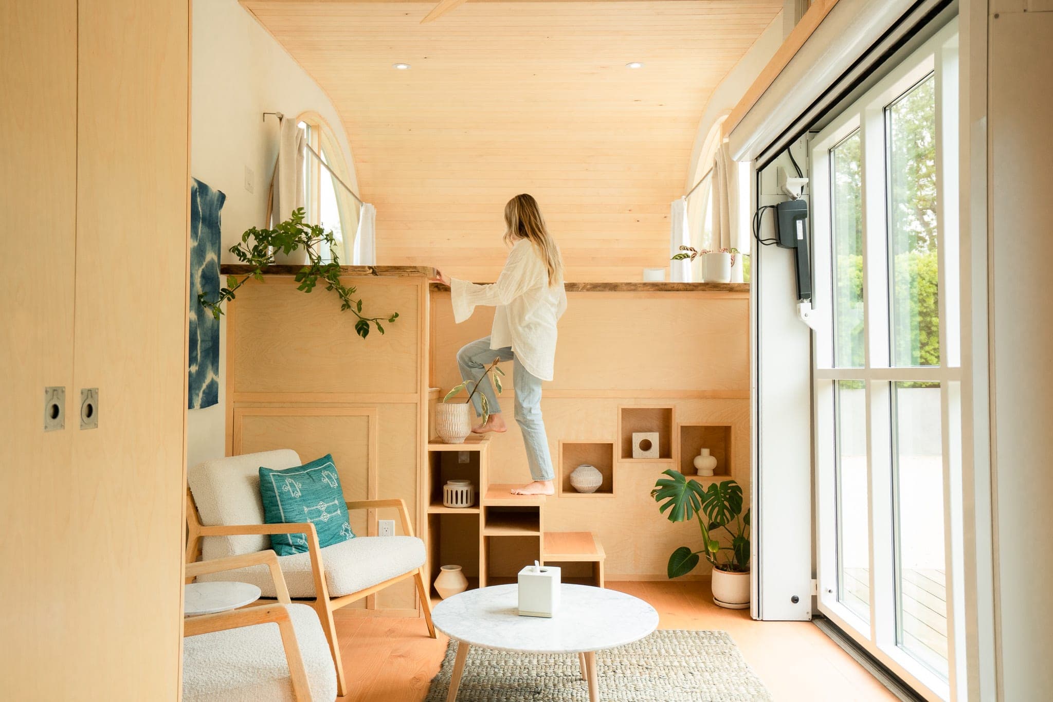 Woman climbing wooden loft stairs in a bright tiny-home interior with plants and a marble coffee table